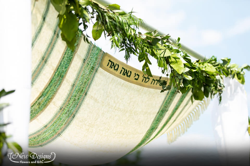 A Chuppah stands against a water front view. Lined with Greenery, and draping white material. With 2 installments of flowers in the middle of each side pole, in various shades of deep and light green and purple.
